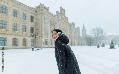A positive Hispanic woman in a coat walks in a blizzard on the streets of the town, looks at the camera and smiles against the backdrop of a beautiful winter view. © bodnarphoto