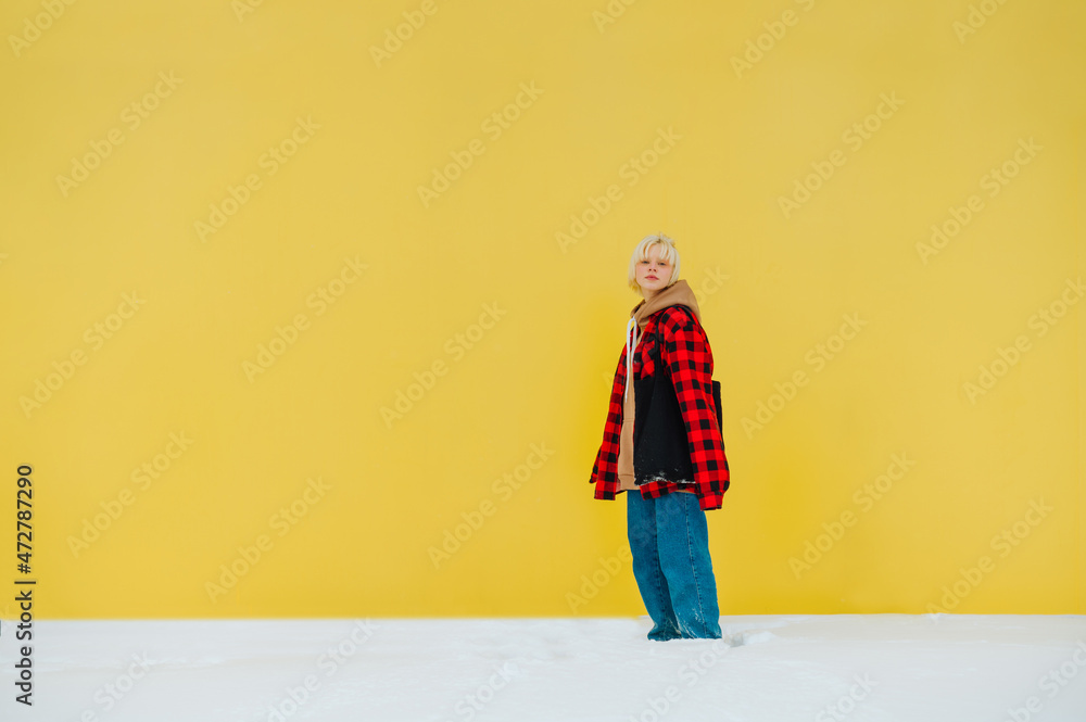 Cute blonde girl stands on the street in winter in the snow on a yellow background and looks at the camera, wears oversized clothes.