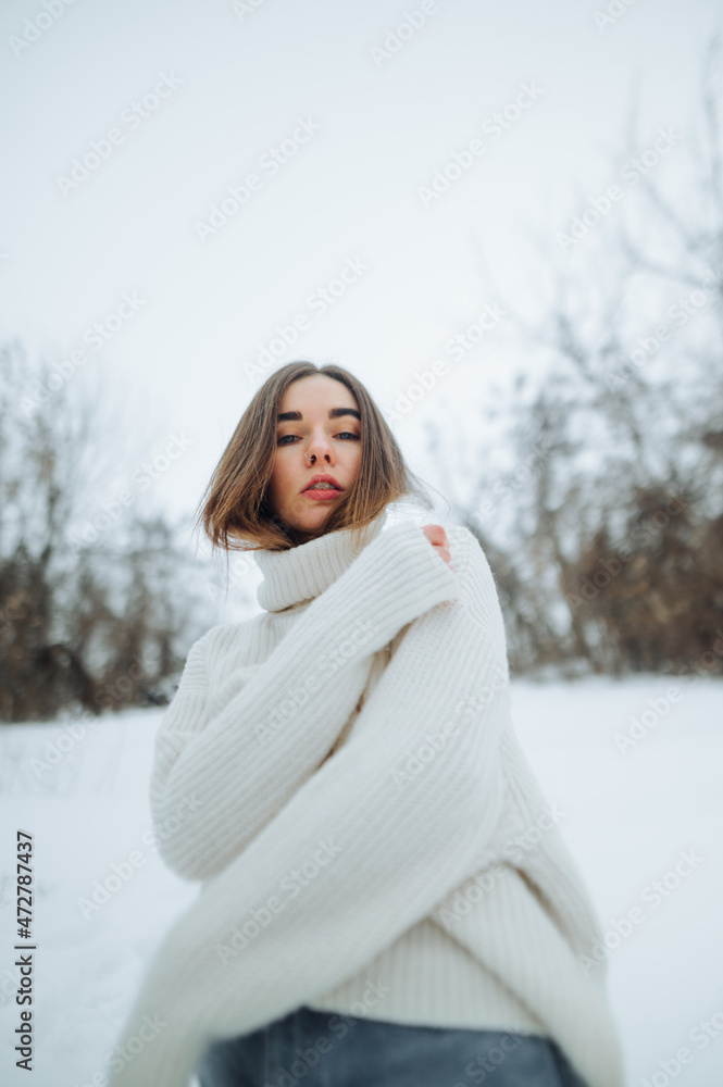 Stylish lady in a white knitted sweater posing for the camera on a snowy street in the winter season, looking at the camera. Vertical. Stylish lady in a white knitted sweater posing for the camera on a snowy street in the winter season, looking at the camera. Vertical.
