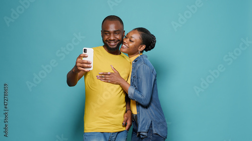 Couple taking selfies with smartphone in front of studio camera. Man and woman making silly faecs while they take pictures together on mobile phone. Casual people enjoying leisure activity photo