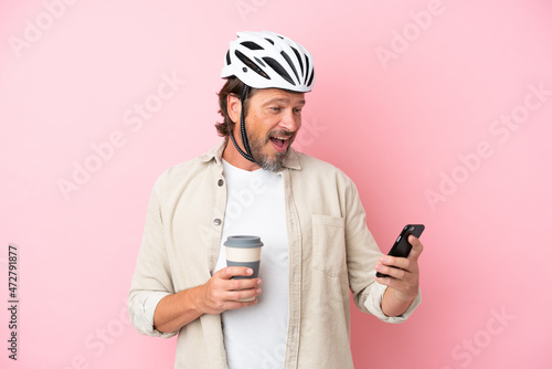 Senior dutch man with bike helmet isolated on pink background holding coffee to take away and a mobile