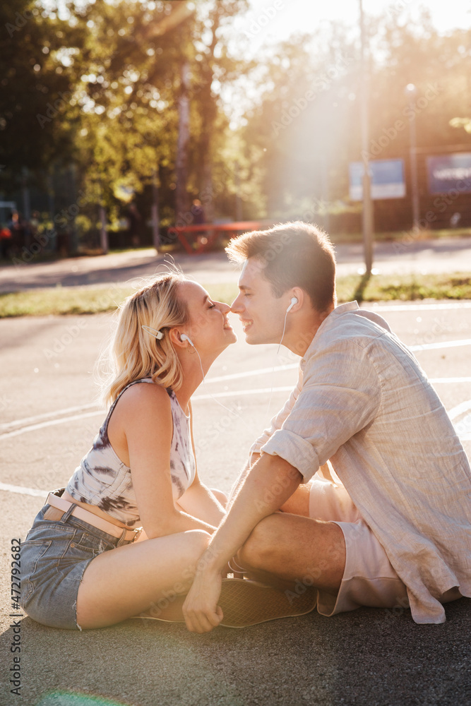 Naklejka premium summer holidays, technology and people concept - happy couple with earphones listening to music and touching noses on basketball playground