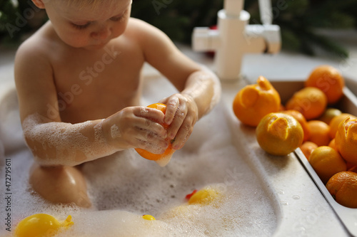 A blonde-haired baby is bathing in the sink in the kitchen against the background of a Christmas tree with garlands and oranges photo
