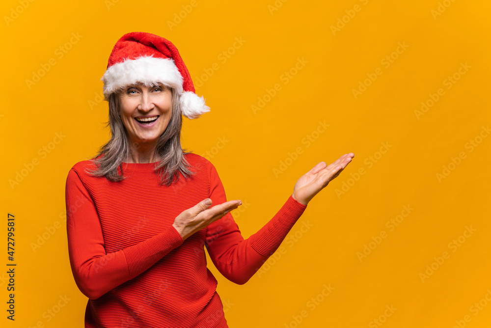 Cheerful and happy senior woman in santa hat points with palms of hands to the empty space, excited mature lady happy to presenting novelty, copy space, mockup, isolated on the yellow Cheerful and happy senior woman in santa hat points with palms of hands to the empty space, excited mature lady happy to presenting novelty, copy space, mockup, isolated on the yellow