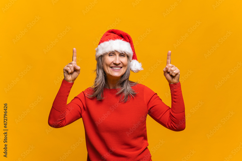 Incredible promotion. Senior woman in santa hat points finger up at empty copy space isolated on yellow, middle-aged lady draws attention to New Year advertising, holiday sales Incredible promotion. Senior woman in santa hat points finger up at empty copy space isolated on yellow, middle-aged lady draws attention to New Year advertising, holiday sales