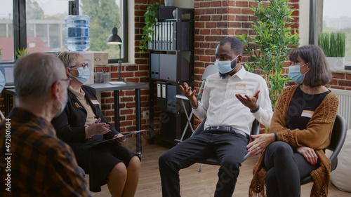 African american man talking to group of people at aa therapy meeting about addiction during pandemic. Person with face mask sharing mental health issues with patients and psychotherapist. photo