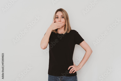 young european woman in black t-shirt while covering her mouth with hand. On white background © Natalya