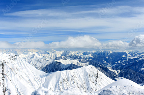 View of snow-covered mountain peaks over which the clouds are floating. In the background is a blue cloudy sky.