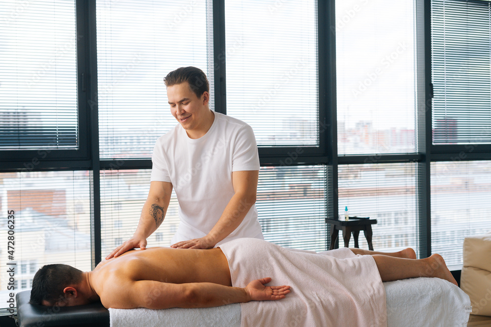 Side wide shot of cheerful male masseur with strong tattooed hands massaging back and shoulder of muscular sports man lying on stomach at massage table. Concept of healthy lifestyle and rest. Side wide shot of cheerful male masseur with strong tattooed hands massaging back and shoulder of muscular sports man lying on stomach at massage table. Concept of healthy lifestyle and rest.