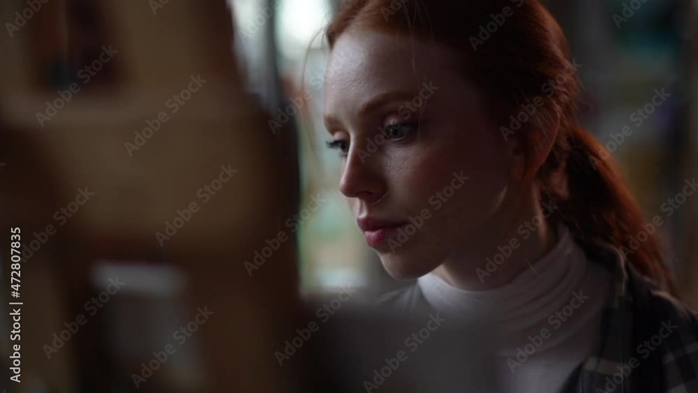 Close-up tracking shot of concentrated young female artist sitting in front of canvas at easel in modern dark art studio making strokes with paintbrush, creating artwork. Shooting in slow motion.