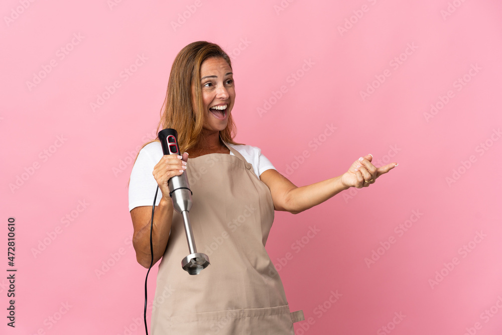 Middle age woman using hand blender isolated on pink background pointing finger to the side and presenting a product Middle age woman using hand blender isolated on pink background pointing finger to the side and presenting a product