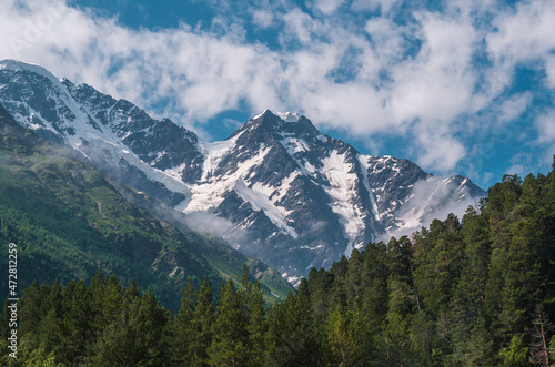 Snowy mountain peaks in sunny summer day with the blue sky and clouds in background, green grass in front. Beautiful landscape of Caucasus mountains.