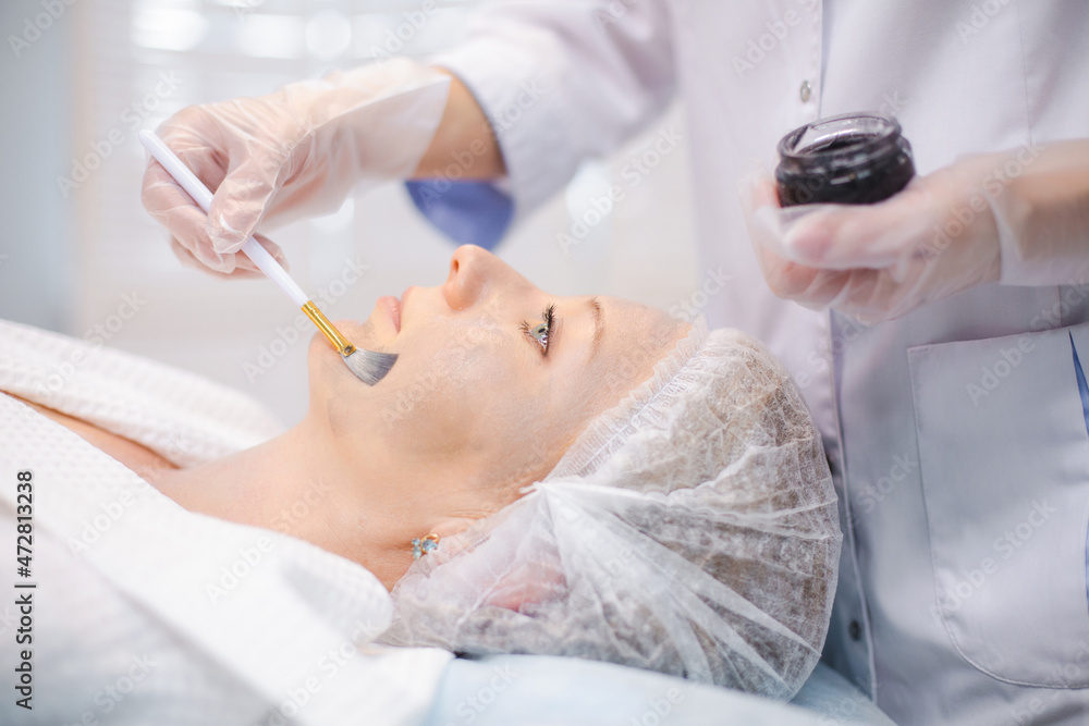 Side view of hands of cosmetology specialist applying carbon nanogel on young woman patient face using brush, getting ready for carbon peeling procedures Side view of hands of cosmetology specialist applying carbon nanogel on young woman patient face using brush, getting ready for carbon peeling procedures