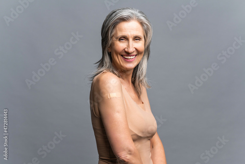 Smiling senior gray-haired woman with a medical patch after vaccination isolated on gray background  protecting hand with bandage after injection. Healthcare and medicine