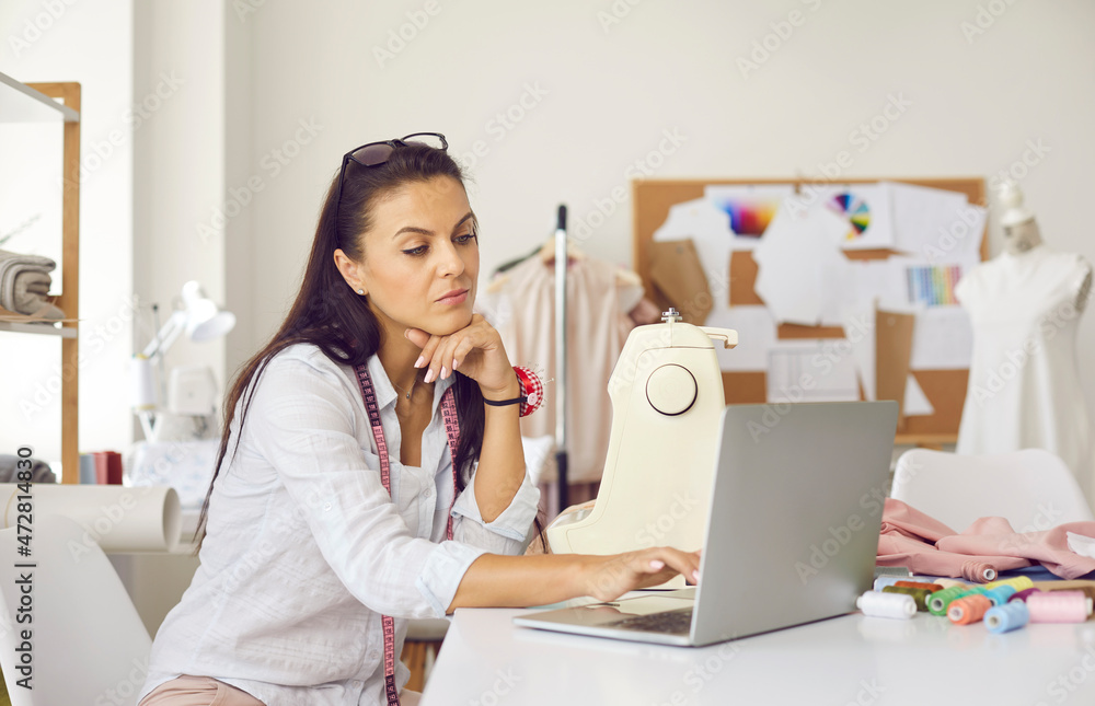 Young Caucasian woman designer work on computer in fashion atelier design styles online on device. Millennial female tailor or seamstress use laptop consult client. Dressmaking, hobby concept. Young Caucasian woman designer work on computer in fashion atelier design styles online on device. Millennial female tailor or seamstress use laptop consult client. Dressmaking, hobby concept.