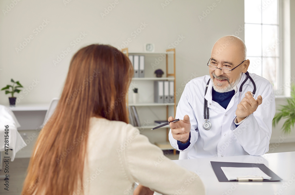 Obraz premium Happy, friendly, smiling senior male doctor giving recommendations to female patient. Cheerful adult man sitting at work desk and talking to young woman who came to hospital for medical consultation