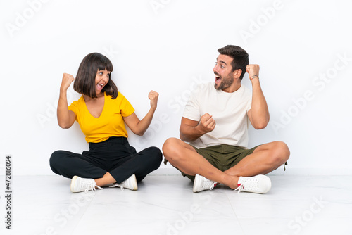 Young couple sitting on the floor isolated on white background celebrating a victory in winner position