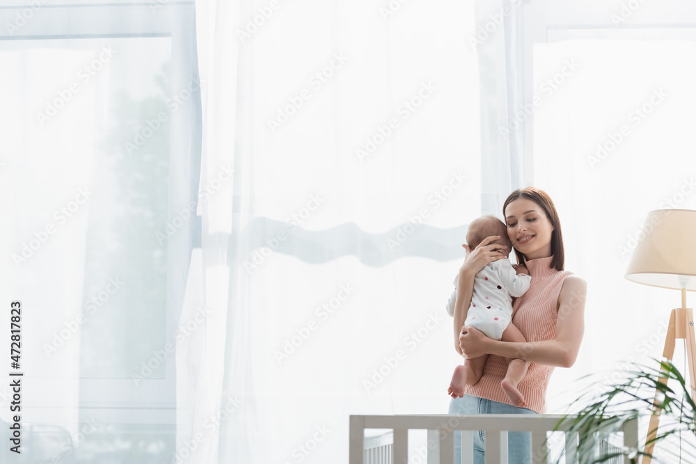 smiling brunette woman with closed eyes embracing toddler son near crib and white curtain. smiling brunette woman with closed eyes embracing toddler son near crib and white curtain.