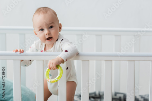 little boy holding rattle ring while standing in crib and looking at camera.