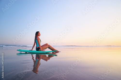 Surfing and vacation. Holiday on the beach. Relaxed young woman sitting on the sand with surf board enjoying sunset.