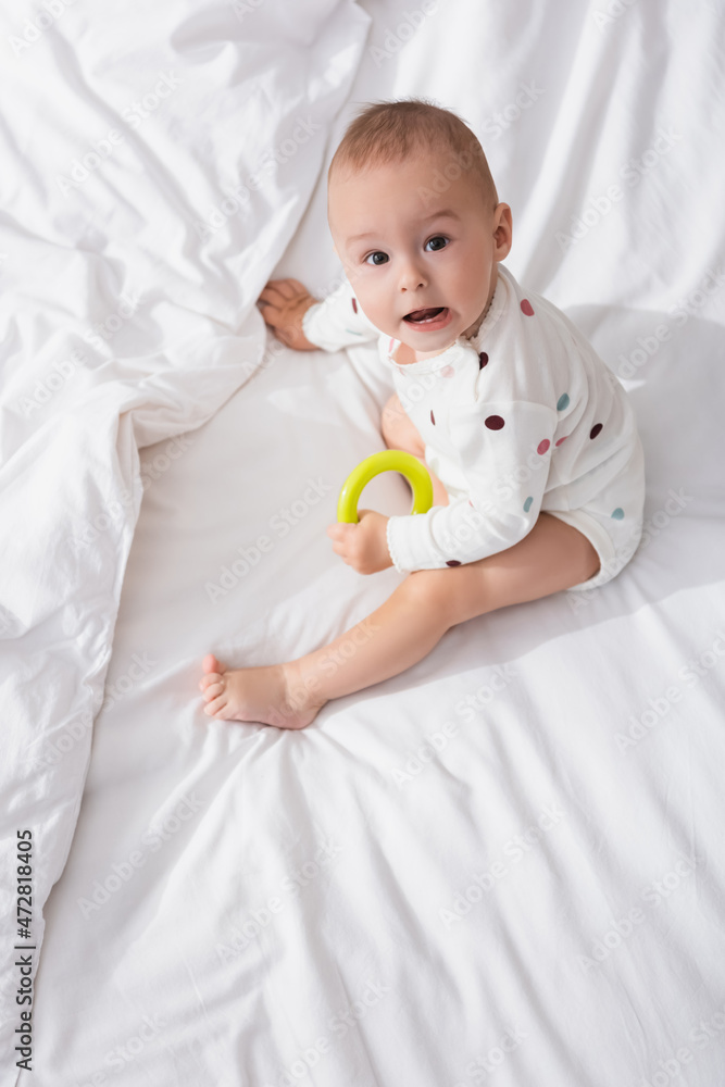 overhead view of little boy with rattle ring looking at camera while sitting on white bedding. overhead view of little boy with rattle ring looking at camera while sitting on white bedding.