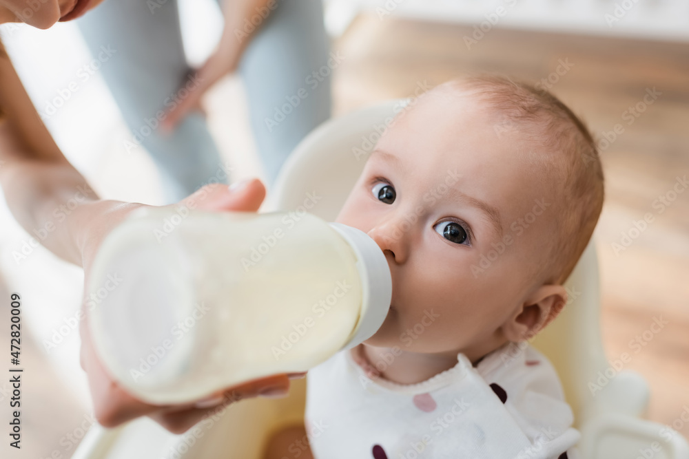 high angle view of toddler boy drinking milk from baby bottle near blurred mom. high angle view of toddler boy drinking milk from baby bottle near blurred mom.