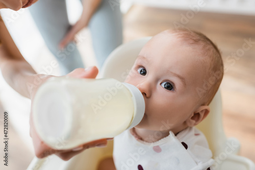 high angle view of toddler boy drinking milk from baby bottle near blurred mom. photo