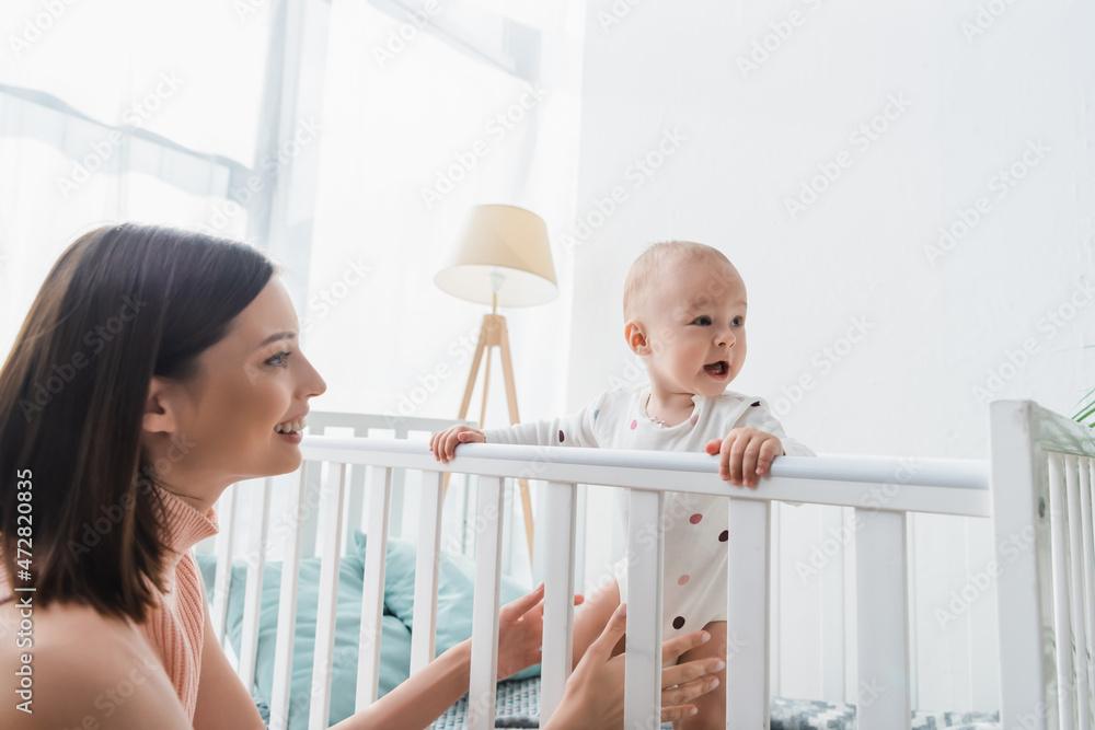 smiling woman near toddler kid standing in crib with open mouth. smiling woman near toddler kid standing in crib with open mouth.