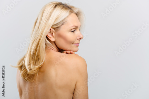 Gorgeous mature blonde middle-aged woman with naked shoulders, looks down and smiling over grey background. Anti-aging treatment and care. Spa and relax, cosmetology procedure