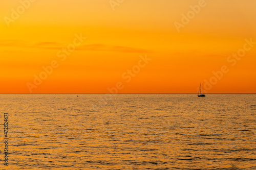 Orange and yellow sunset on the sea with a sailboat. Minimalistic photo of a silhouette of sailing boat on the sea during dusk. Golden sundown and sailing boat silhouette on right.