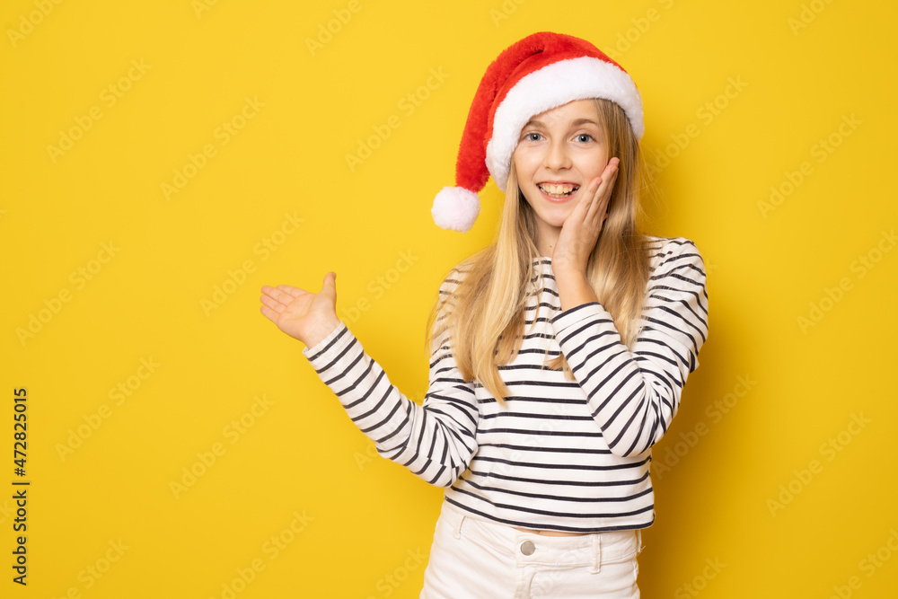 Smiling girl in santa hat showing product with hand isolated over yellow background. Smiling girl in santa hat showing product with hand isolated over yellow background.