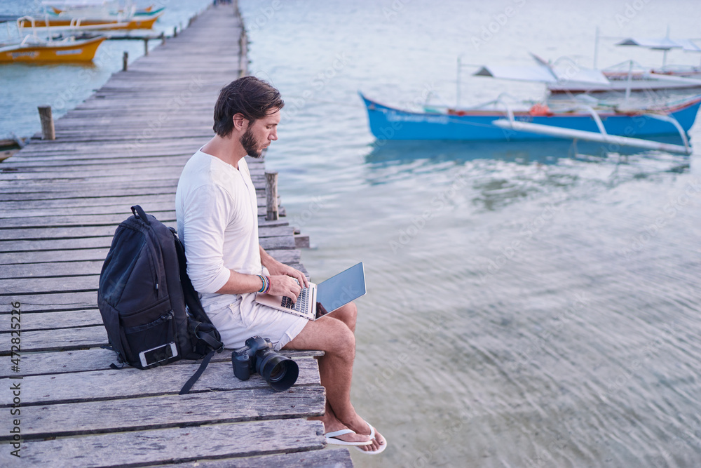 Work and travel. Young man with rucksack using laptop computer sitting on wooden fishing pier with beautiful tropical sea view. Work and travel. Young man with rucksack using laptop computer sitting on wooden fishing pier with beautiful tropical sea view.