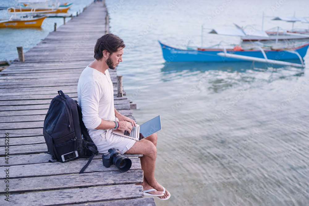 Work and travel. Young man with rucksack using laptop computer sitting on wooden fishing pier with beautiful tropical sea view.