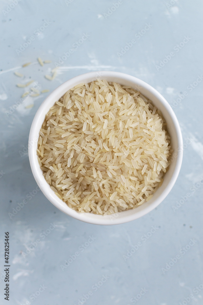 White milled long grain rice in a white bowl on a gray background. Vegetarian food concept. Vertical orientation. Rice dishes are cooked in all countries of the world. Popular cereal. Top view. White milled long grain rice in a white bowl on a gray background. Vegetarian food concept. Vertical orientation. Rice dishes are cooked in all countries of the world. Popular cereal. Top view.
