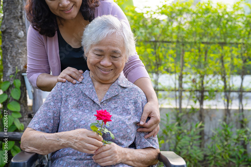 Caregiver daughter hug and help  Asian senior or elderly old lady woman holding red rose on wheelchair in park.