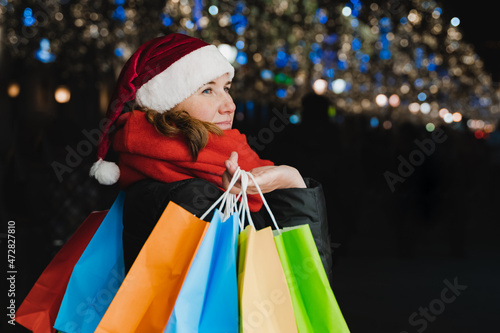 Christmas shopping of young beautiful woman. Colorful bright paper bags with gifts,presents.Walking on market street in city.Garlands,lights.Stylish look, jacket, santa claus hat.New year celebration