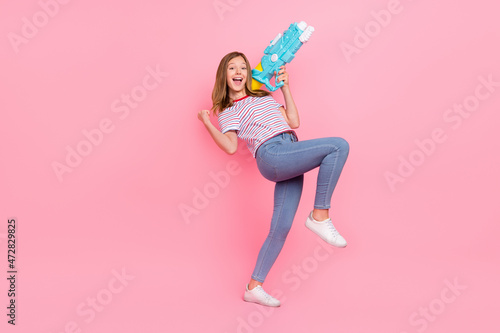 Full size photo of hooray small blond girl hold gun wear t-shirt jeans sneakers isolated on pink background
