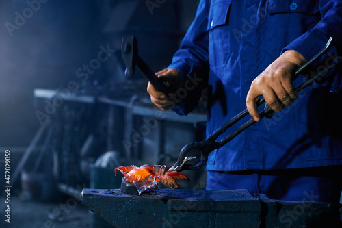 Side view of worker in dark blue suit holding hammer and creating metal for modern fence in smithy. Concept of process working with special modern heating equipments and tools. 