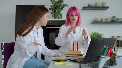 Concentrated pretty pink haired researcher and lovely female student with congenital physical anomaly of hands wearing lab coats, carrying out scientific experiments with test tubes indoors. photo