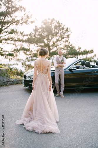 Bride walks along the road to groom standing near the convertible © Nadtochiy