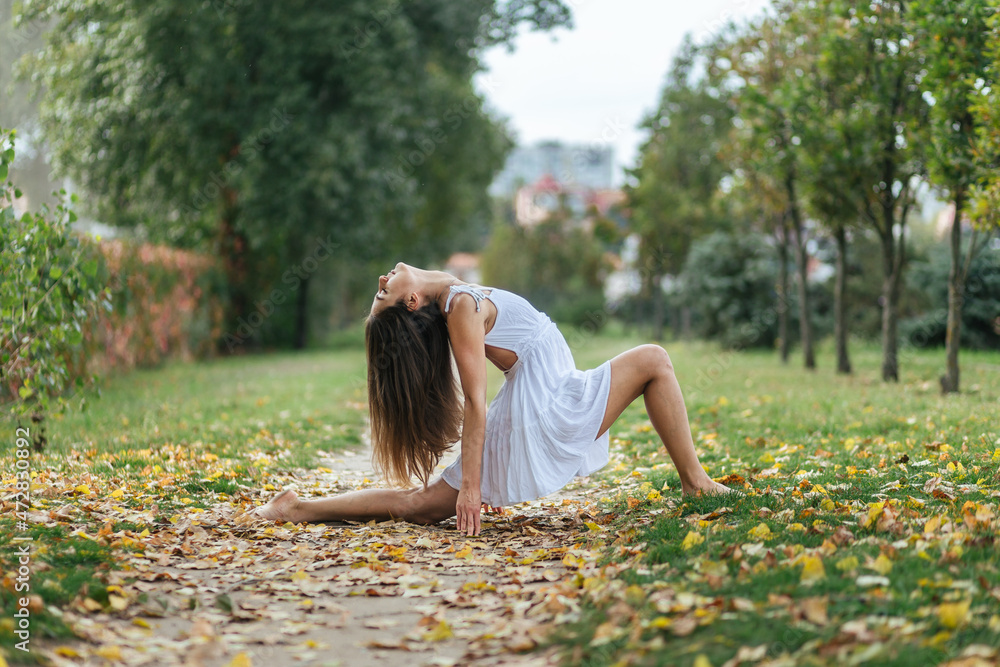 Fototapeta premium beautiful girl in white dress doing yoga poses in the autumn park, balance, fitness, stretching and relaxation