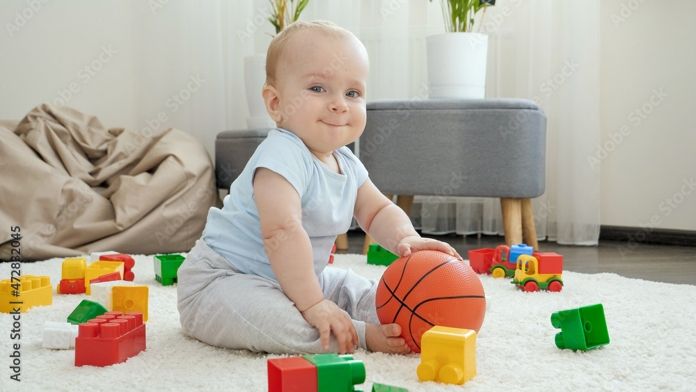 Cute smiling baby boy sitting on carpet and holding basketball ball Cute smiling baby boy sitting on carpet and holding basketball ball