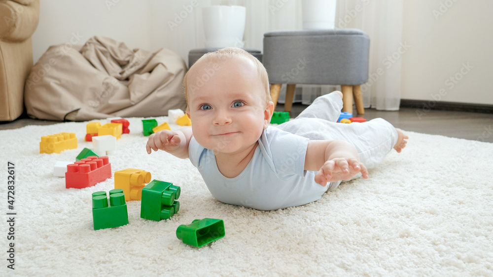 Happy cheerfull baby boy lying on floor next to colorful toys, bricks and blocks at playroom. Concept of children development, sports, education and creativity at home Happy cheerfull baby boy lying on floor next to colorful toys, bricks and blocks at playroom. Concept of children development, sports, education and creativity at home