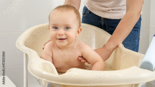 Happy laughing baby boy sitting in plastic bath and splashing water. Concept of children hygiene, healthcare and parenting.