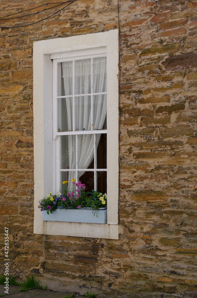 A white-framed window with a flower box set into a brown stone wall.