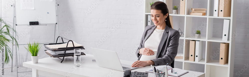 © LIGHTFIELD STUDIOS - happy pregnant businesswoman looking at laptop in office, banner.