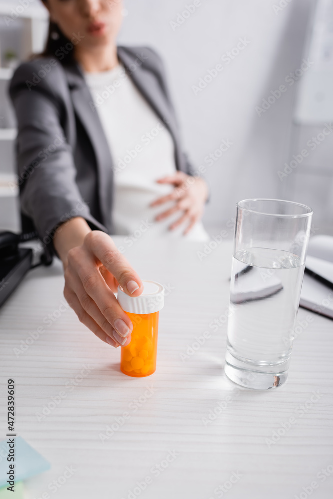 cropped view of blurred pregnant woman reaching jar with pills near glass of water on desk. cropped view of blurred pregnant woman reaching jar with pills near glass of water on desk.