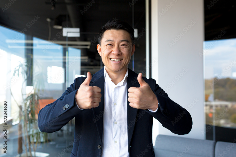 Asian happy man looking at camera showing thumbs up and ok sign, businessman smiling in modern office Asian happy man looking at camera showing thumbs up and ok sign, businessman smiling in modern office