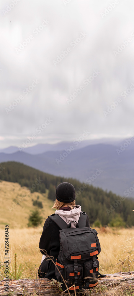 A woman with a backpack sitting on the slope on a fallen tree and enjoying the scenic view. Hiking and climbing to the top of the mountain.