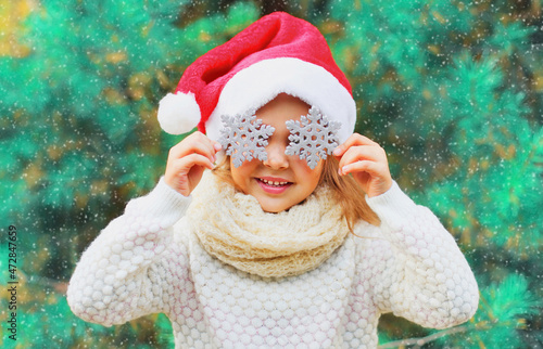 Christmas portrait of child in santa red hat covering her eyes with a snowflakes toy over a tree